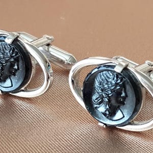 May include: A pair of silver cufflinks with oval black cameos depicting a woman's profile. The cameos are set in a silver oval frame. The cufflinks have a classic, elegant design.