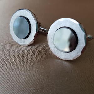 May include: A pair of silver cufflinks with a faceted edge design. Each cufflink has a circular, iridescent, grey-blue centre stone surrounded by a black border. The cufflinks are connected by a small, silver bar.