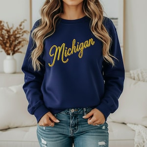 May include: A navy blue long-sleeve crewneck sweatshirt featuring the word Michigan written in a yellow cursive script font with a thin white outline. The sweatshirt has a ribbed collar, cuffs, and hem, and is paired with blue denim jeans.