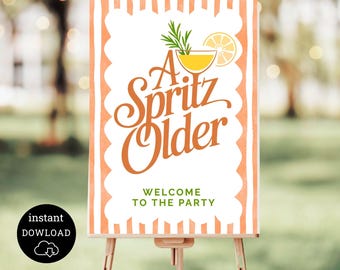 May include: A Spritz Older party welcome sign on a wooden easel. The sign has a white background with orange scalloped edges and vertical stripes. The text "A Spritz Older" is in orange script, with a cocktail illustration and the words "Welcome to the Party".