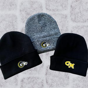 May include: Three beanies in a square composition. Two black beanies and one gray beanie are displayed. Each beanie has a yellow and white embroidered logo. The logos include a stylized wolf and the letters "GM".