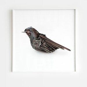 May include: A metal sculpture of a bird with brown and silver wings. The bird is perched on a white surface and is framed in a white frame.