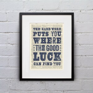 May include: Framed print with the text "THE HARD WORK PUTS YOU WHERE THE GOOD LUCK CAN FIND YOU" in navy blue. The text is set against a vintage dictionary page background, framed in black with a white mat.