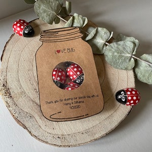 Mason jar shaped wedding favours with a circle cutout in the centre showing bug shaped sweets. Text Love Bug at they top and couples name and date at the bottom.