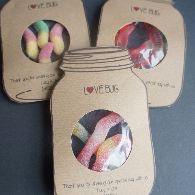 Mason jar shaped wedding favours with a circle cutout in the centre showing bug shaped sweets. Text Love Bug at they top and couples name and date at the bottom.