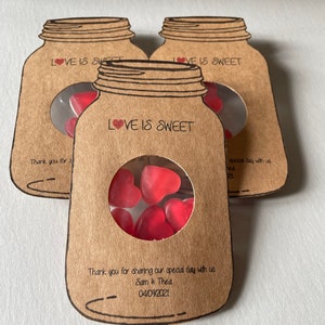 Mason Jar shaped favours made out of natural kraft card stock with a circle window in the centre displaying heart shaped sweets. Love is sweet text at the top of the favour and couples names and wedding date at the bottom. Styled as a set of three