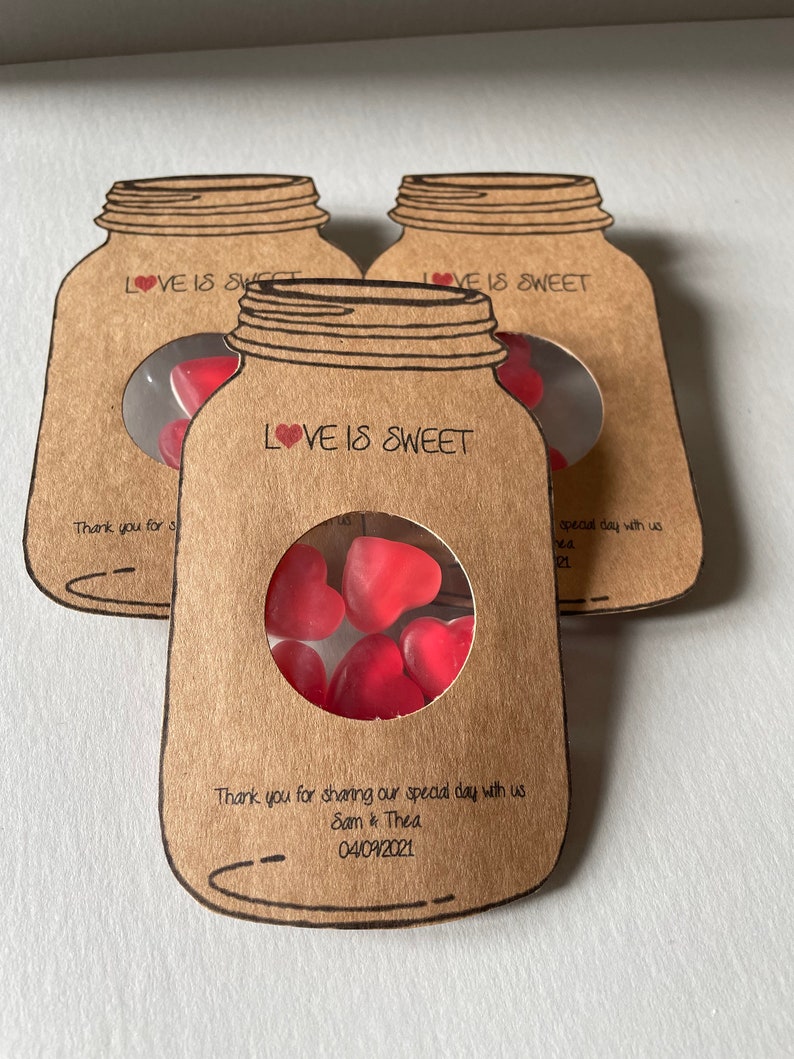 Mason Jar shaped favours made out of natural kraft card stock with a circle window in the centre displaying heart shaped sweets. Love is sweet text at the top of the favour and couples names and wedding date at the bottom. Styled as a set of three