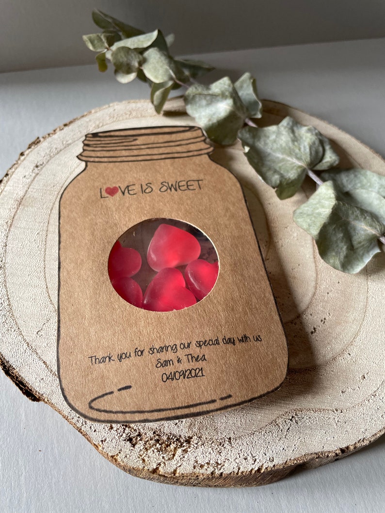 Mason Jar shaped favours made out of natural kraft card stock with a circle window in the centre displaying heart shaped sweets. Love is sweet text at the top of the favour and couples names and wedding date at the bottom.