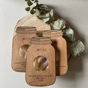 Mason jar shaped wedding favours with a circle cutout in the centre showing bug shaped sweets. Text Love Bug at they top and couples name and date at the bottom. Styled as a set of three