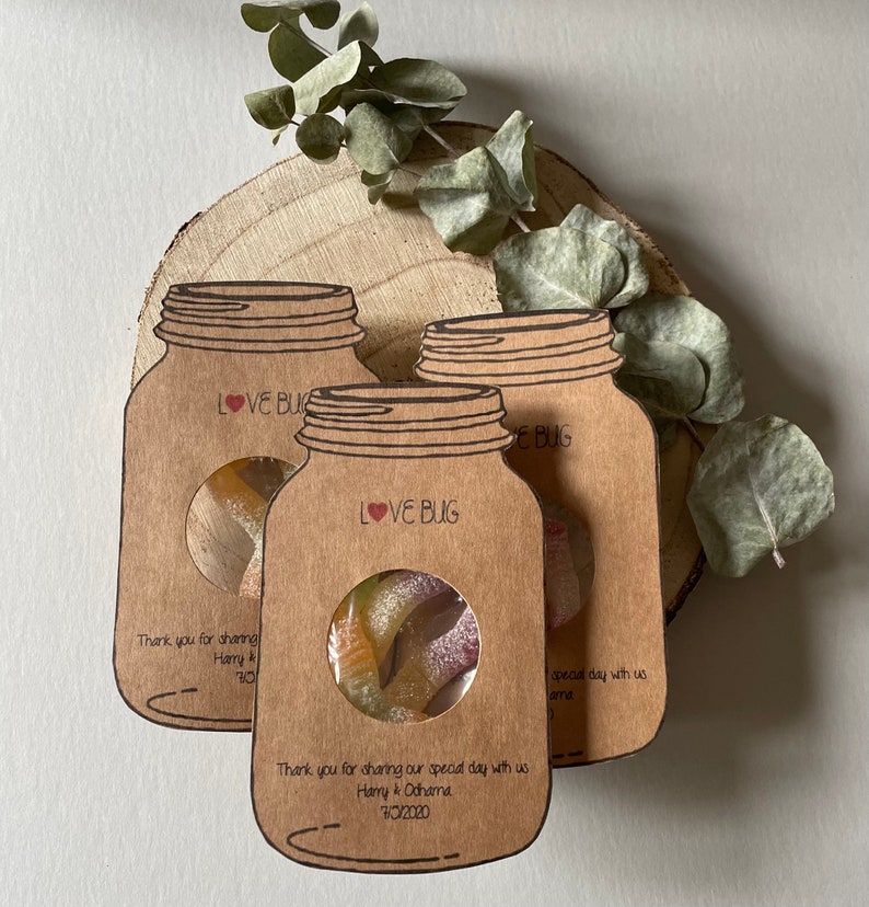 Mason jar shaped wedding favours with a circle cutout in the centre showing bug shaped sweets. Text Love Bug at they top and couples name and date at the bottom. Styled as a set of three