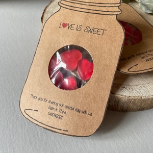 Mason Jar shaped favours made out of natural kraft card stock with a circle window in the centre displaying heart shaped sweets. Love is sweet text at the top of the favour and couples names and wedding date at the bottom.