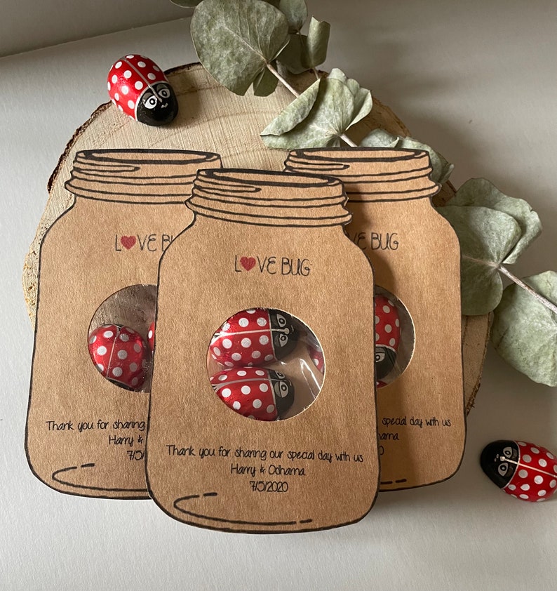 Mason jar shaped wedding favours with a circle cutout in the centre showing bug shaped sweets. Text Love Bug at they top and couples name and date at the bottom. Styled as a set of three