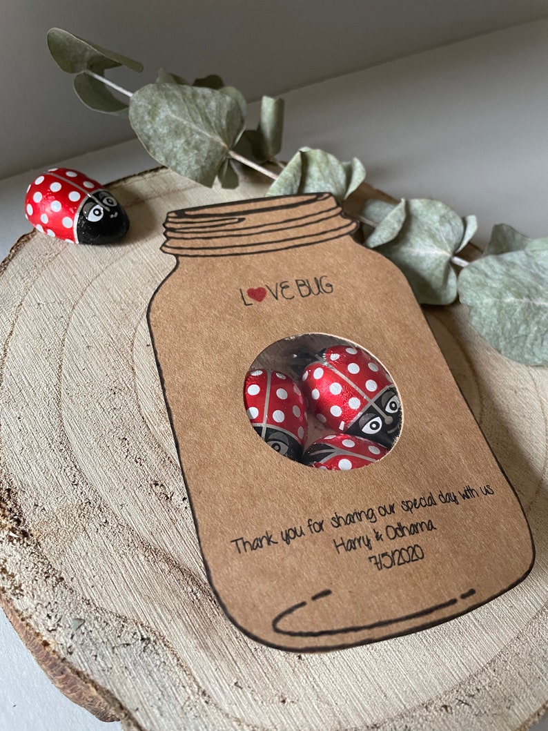 Mason jar shaped wedding favours with a circle cutout in the centre showing bug shaped sweets. Text Love Bug at they top and couples name and date at the bottom. Styled on a natural wood slice