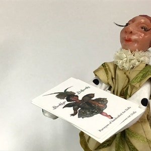 Puede incluir: Una muñeca con un vestido verde y dorado con lunares negros sostiene una tarjeta blanca con el texto "Brooklyn Dollworks" y "Purveyors of handcrafted Art Dolls".