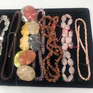 May include: A collection of various gemstone beads and strands, including clear, brown, pink, and tan colors. The beads are arranged in a black tray, showcasing different shapes and sizes, suitable for jewelry making.