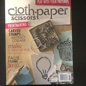 May include: A magazine cover for "Cloth Paper Scissors" with the title text in large, stylized letters. The cover features printmaking examples, including carved stamps and a lightbulb design. The magazine promotes collage and mixed media art.