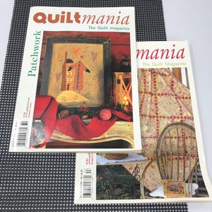 May include: Two copies of "Quiltmania" magazine are displayed. The covers feature quilt-themed imagery, including a framed patchwork design and a quilt draped over a chair. The magazine's title is prominently displayed in red and white.