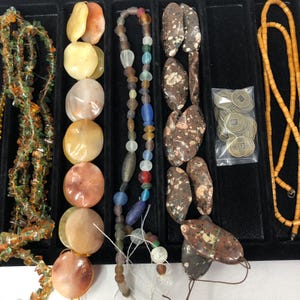 May include: Assortment of beads and jewelry components in a black tray. Includes strands of colorful glass beads, polished stone beads in shades of orange and brown, and small metal coins. A variety of shapes and sizes are visible.