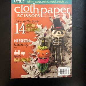 May include: A magazine cover with an orange background features the text "cloth paper scissors" and "Day of the Dead." The cover showcases Halloween-themed art projects, including a pumpkin and a skeleton doll.