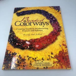May include: A book titled "Beaded Colorways" with a yellow cover. The cover features a beaded necklace design in shades of purple, red, and orange. The book's title and author's name are visible. The text "Includes Custom Color Wheels" is also present.