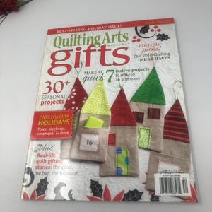 May include: A magazine cover for "Quilting Arts Gifts" features a holiday theme with colorful fabric house ornaments. The cover includes text about seasonal projects, patchwork holidays, and real-life quilt gifting stories.
