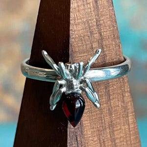May include: A silver-toned ring featuring a detailed spider design with a teardrop-shaped, dark red gemstone. The spider's body is the gemstone, and the legs extend outward. The ring is displayed on a wooden stand.