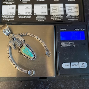 May include: A silver pendant with a turquoise stone is on a digital scale, showing 26.1 grams. The pendant has a horseshoe design with floral and geometric patterns. The scale's instructions are visible in the background.