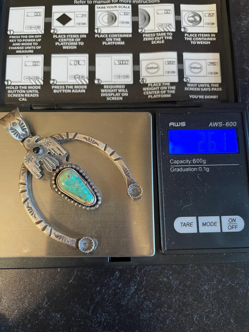 May include: A silver pendant with a turquoise stone is on a digital scale, showing 26.1 grams. The pendant has a horseshoe design with floral and geometric patterns. The scale's instructions are visible in the background.