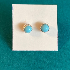 May include: A pair of stud earrings featuring round, smooth, light blue turquoise cabochons set in scalloped silver-toned metal settings. The earrings are mounted on a small, square white display card for presentation and storage.