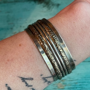 May include: A stack of silver-toned bangles with etched floral designs. The bracelets are worn on a wrist, showcasing the intricate patterns and textures of the jewelry. The bangles vary in width and design, creating a layered effect.