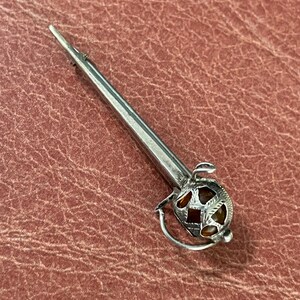 May include: A silver-toned kilt pin in the shape of a sword. The pin features a detailed hilt with a decorative design and a pointed blade. The pin is set against a textured, reddish-brown surface.