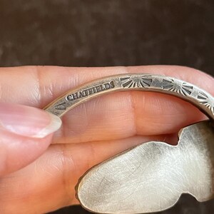 May include: Close-up of a silver bracelet with the engraved text "CHATFIELDS". The bracelet features a sunburst pattern and an organic shape. The metal has a brushed finish. The bracelet is made of silver.