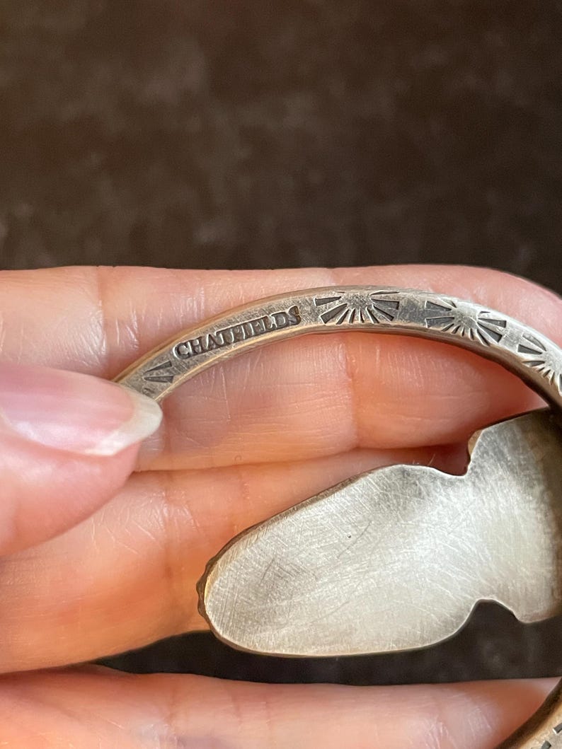 May include: Close-up of a silver bracelet with the engraved text "CHATFIELDS". The bracelet features a sunburst pattern and an organic shape. The metal has a brushed finish. The bracelet is made of silver.
