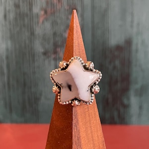 May include: A silver ring with a star-shaped stone. The stone is white with gray marbling. The ring has a decorative border with small floral accents. The ring is displayed on a wooden stand.