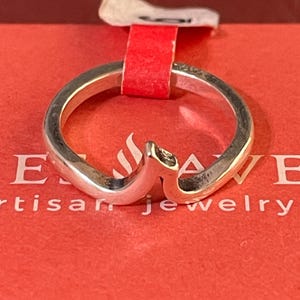 May include: A silver ring with a smooth, polished finish and a distinct V-shaped chevron design at the top. The band features a consistent thickness throughout its circumference, showcasing a clean, metallic luster and a simple, unadorned aesthetic.