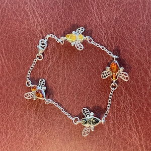 May include: A silver bracelet featuring four bee charms with amber and green-toned accents. The bees have detailed wings and are connected by a delicate chain. The bracelet is displayed on a textured, reddish-brown surface.