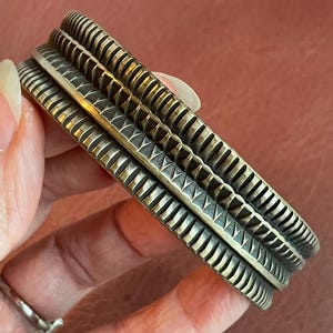 K&ouml;nnte beinhalten: Ein silbernes Armband mit geometrischem Muster. Das Armband besteht aus zwei B&auml;ndern mit einer strukturierten Oberfl&auml;che.