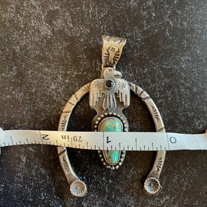 May include: Silver pendant necklace featuring a turquoise stone and black accent. The pendant is approximately 5 cm wide. The design includes an eagle and a horseshoe shape, with decorative details.