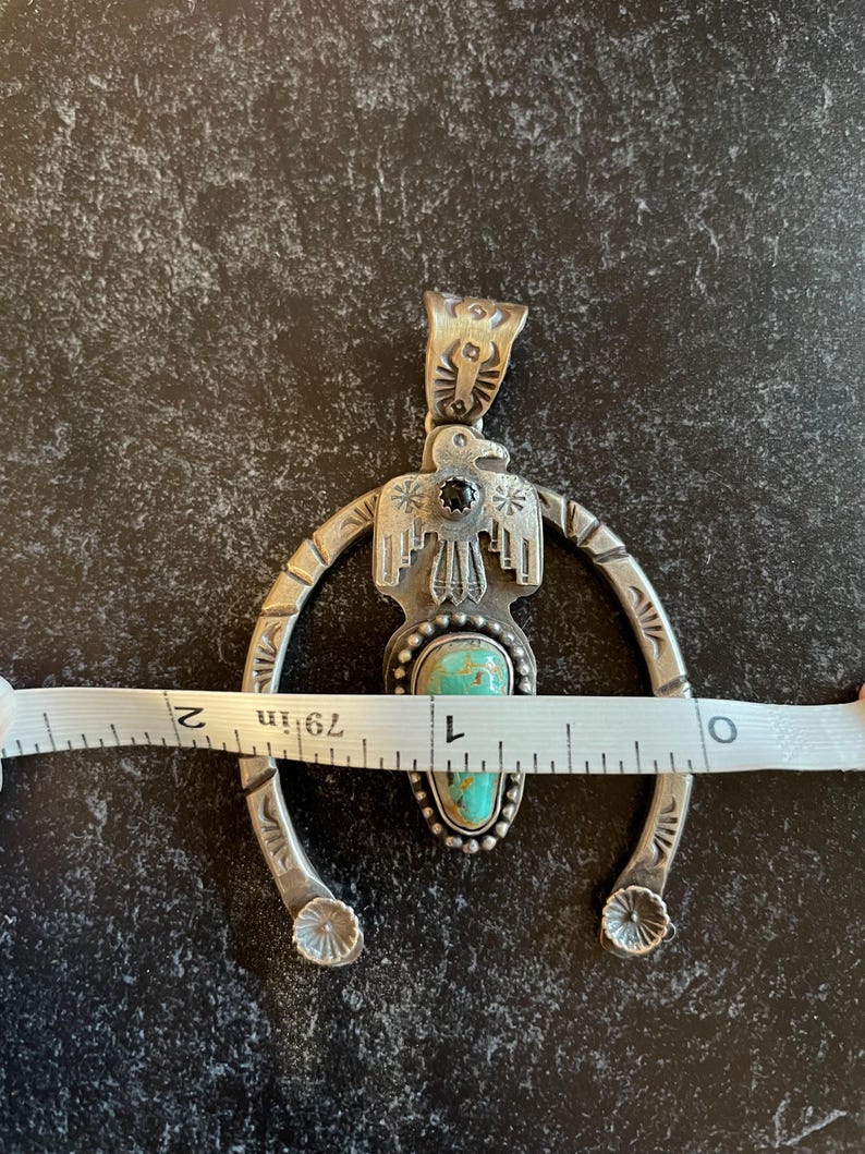 May include: Silver pendant necklace featuring a turquoise stone and black accent. The pendant is approximately 5 cm wide. The design includes an eagle and a horseshoe shape, with decorative details.