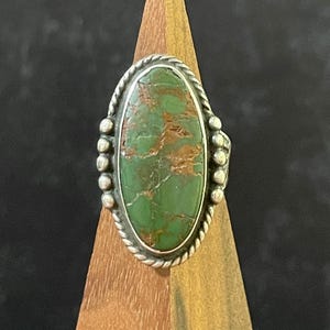 May include: An oval-shaped ring featuring a green stone with brown matrix veining. The stone is set in a silver bezel surrounded by a twisted rope border and accented with small silver spheres on both sides of the setting, mounted on a band.