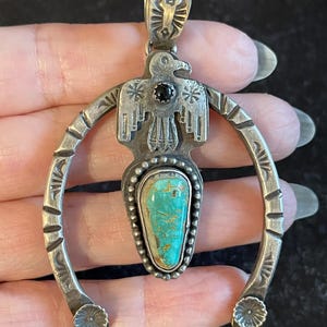 May include: Vintage silver pendant featuring a turquoise stone and a bird design. The pendant has a horseshoe shape with floral accents and a black stone. The turquoise stone is a light blue-green colour.