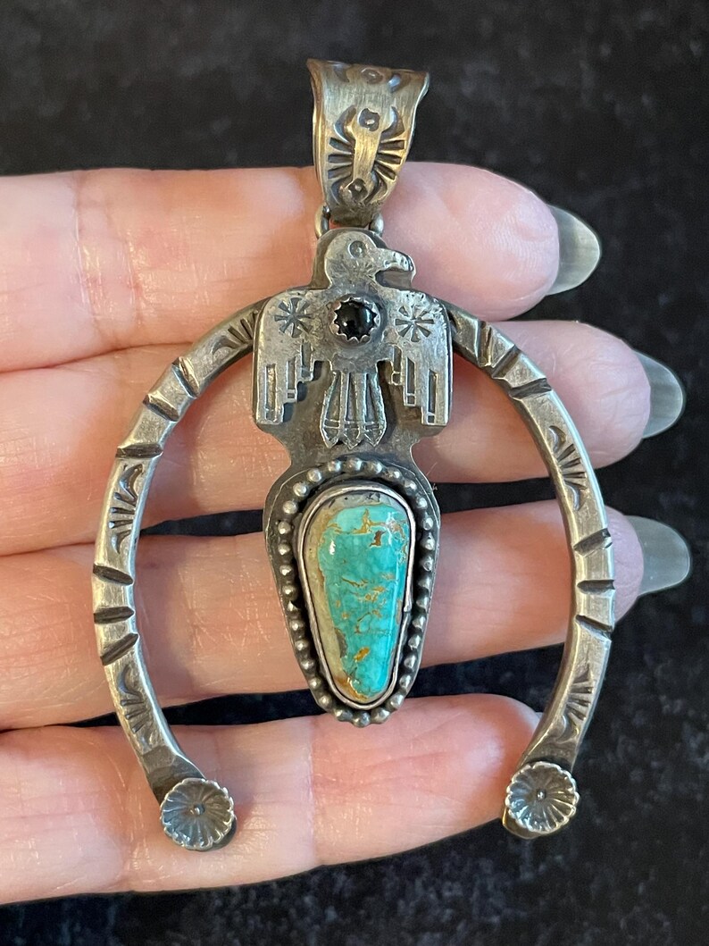 May include: Vintage silver pendant featuring a turquoise stone and a bird design. The pendant has a horseshoe shape with floral accents and a black stone. The turquoise stone is a light blue-green colour.