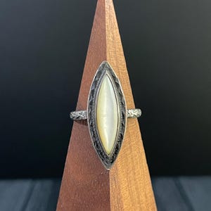 May include: A silver ring with a marquise-shaped, cream-colored stone. The stone is set in a detailed, dark-toned bezel. The band has a textured design. The ring is displayed on a wooden stand against a dark background.
