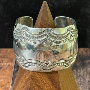 May include: A wide silver cuff bracelet featuring intricate stamped patterns along the top and bottom edges. The polished metal surface displays symmetrical, repeating geometric designs with fine lines and semi-circular motifs encircling the band.
