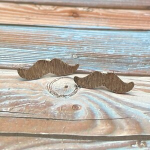 May include: A pair of silver mustache shaped earrings on a wooden background.