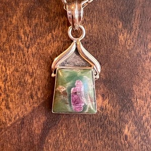 May include: A silver pendant necklace featuring a rectangular green stone with a pink crystal inclusion. The pendant is suspended from a silver chain. The necklace is displayed against a brown wooden background.