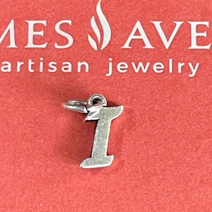 May include: A silver-colored charm in the shape of the letter "I" with a small loop at the top for attachment. The charm is set against a red background with the text "MES AVE artisan jewelry".