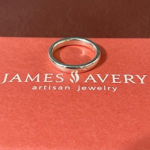 May include: A polished silver band ring resting on a coral-colored box with the white text JAMES AVERY artisan jewelry printed on the surface. The ring features a smooth, rounded exterior surface and a simple, continuous circular design.
