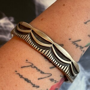 May include: A silver bracelet with a decorative design of repeating curved shapes. The bracelet is worn on a person's wrist.