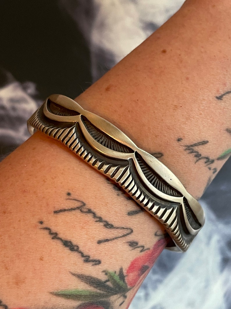 May include: A silver bracelet with a decorative design of repeating curved shapes. The bracelet is worn on a person's wrist.